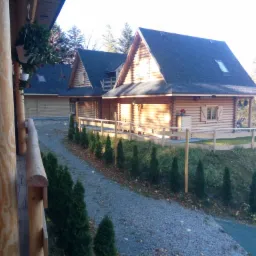 Widok z drewnianego balkonu na dwa domki letniskowe z bali, oświetlone popołudniowym słońcem, z ciemnymi dachami i oknami ze szprosami, otoczone niskimi tujami i żwirową ścieżką.