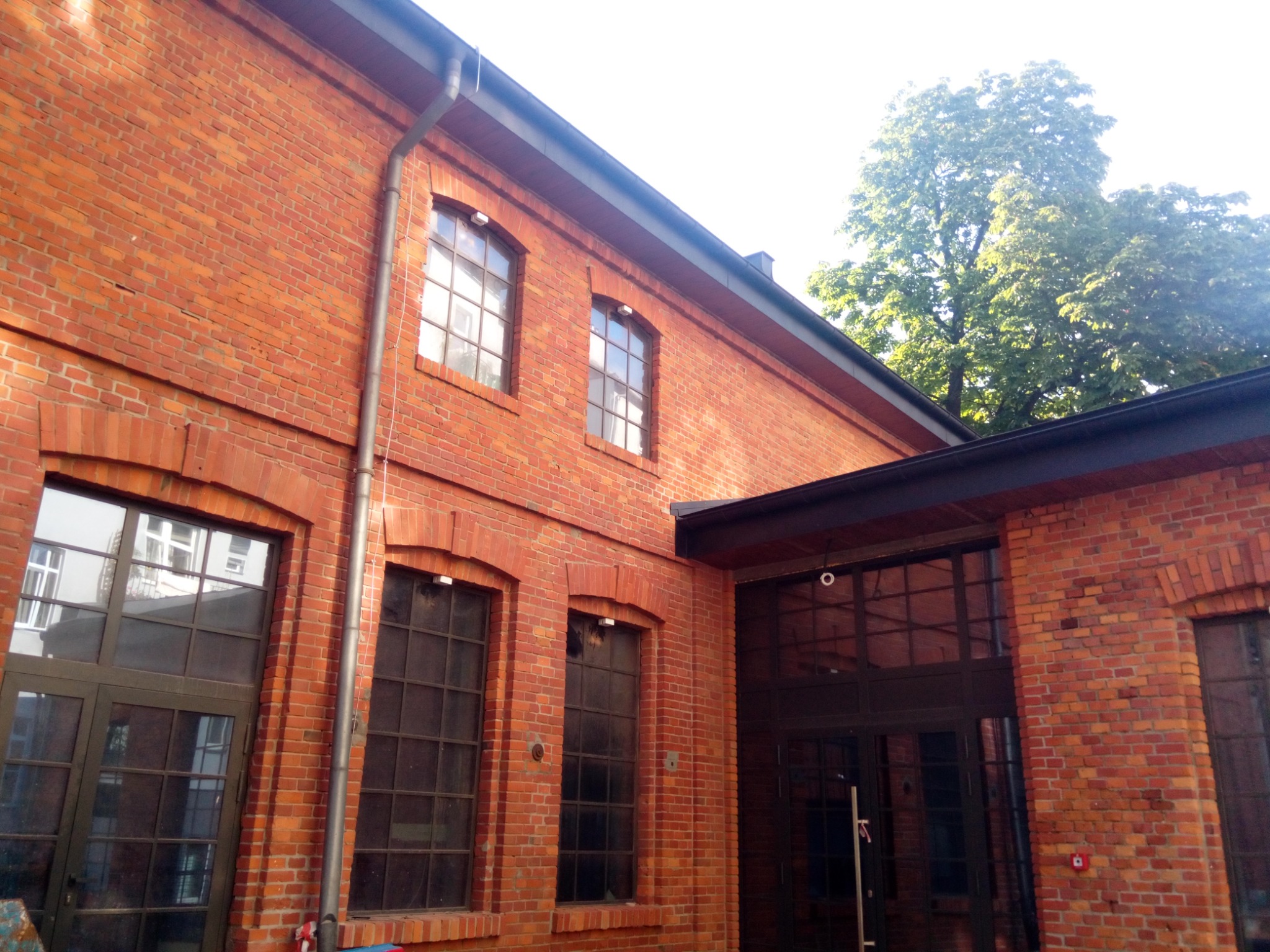 Ceglany budynek z dużymi oknami i drzwiami, widoczne rynny i oświetlenie zewnętrzne, zieleń drzew w tle.