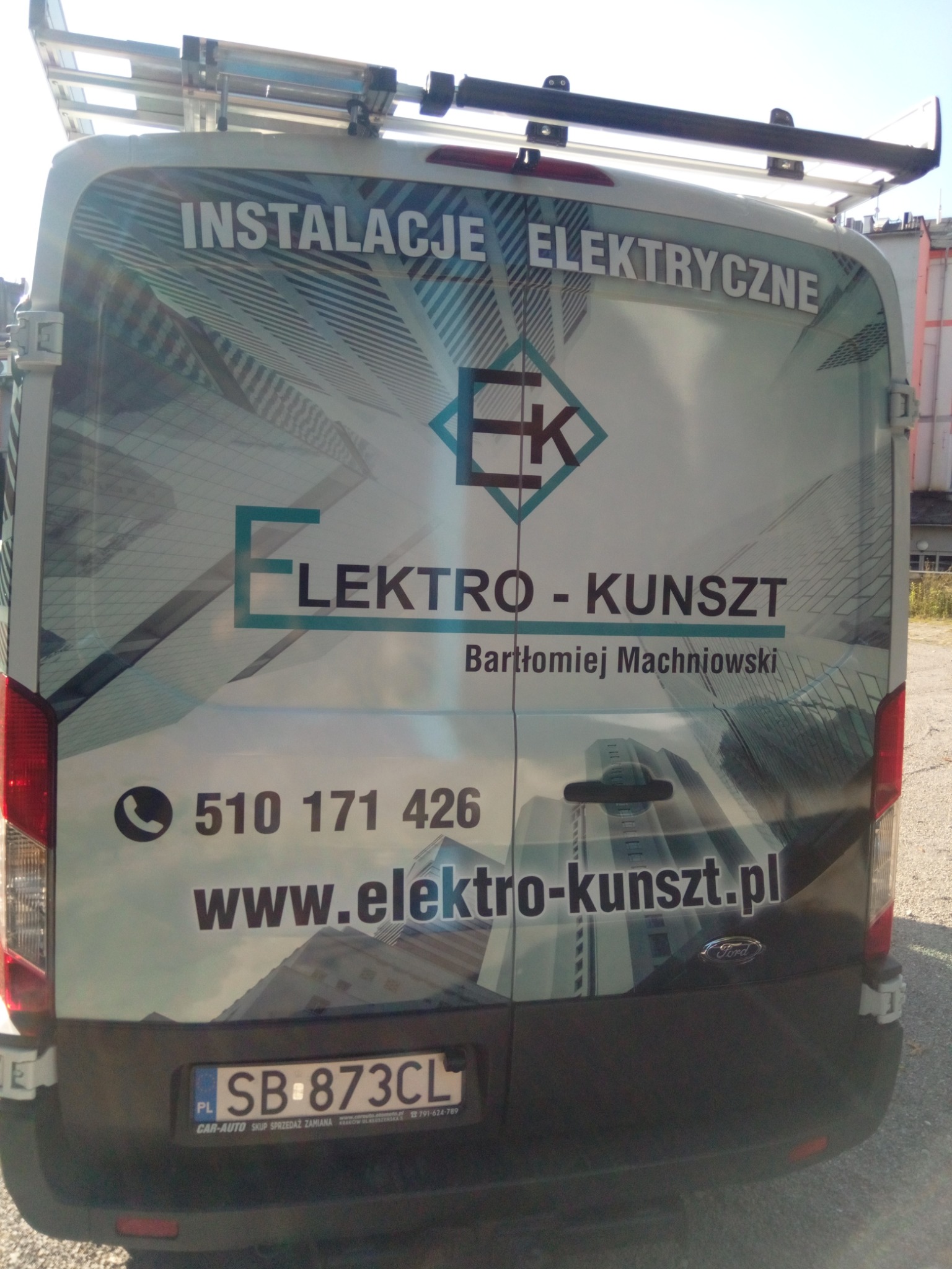 Tył białego samochodu dostawczego z reklamą firmy Elektro-Kunszt, oferującej usługi elektryczne, widoczny numer telefonu i adres strony internetowej na tle grafiki przedstawiającej budynki.