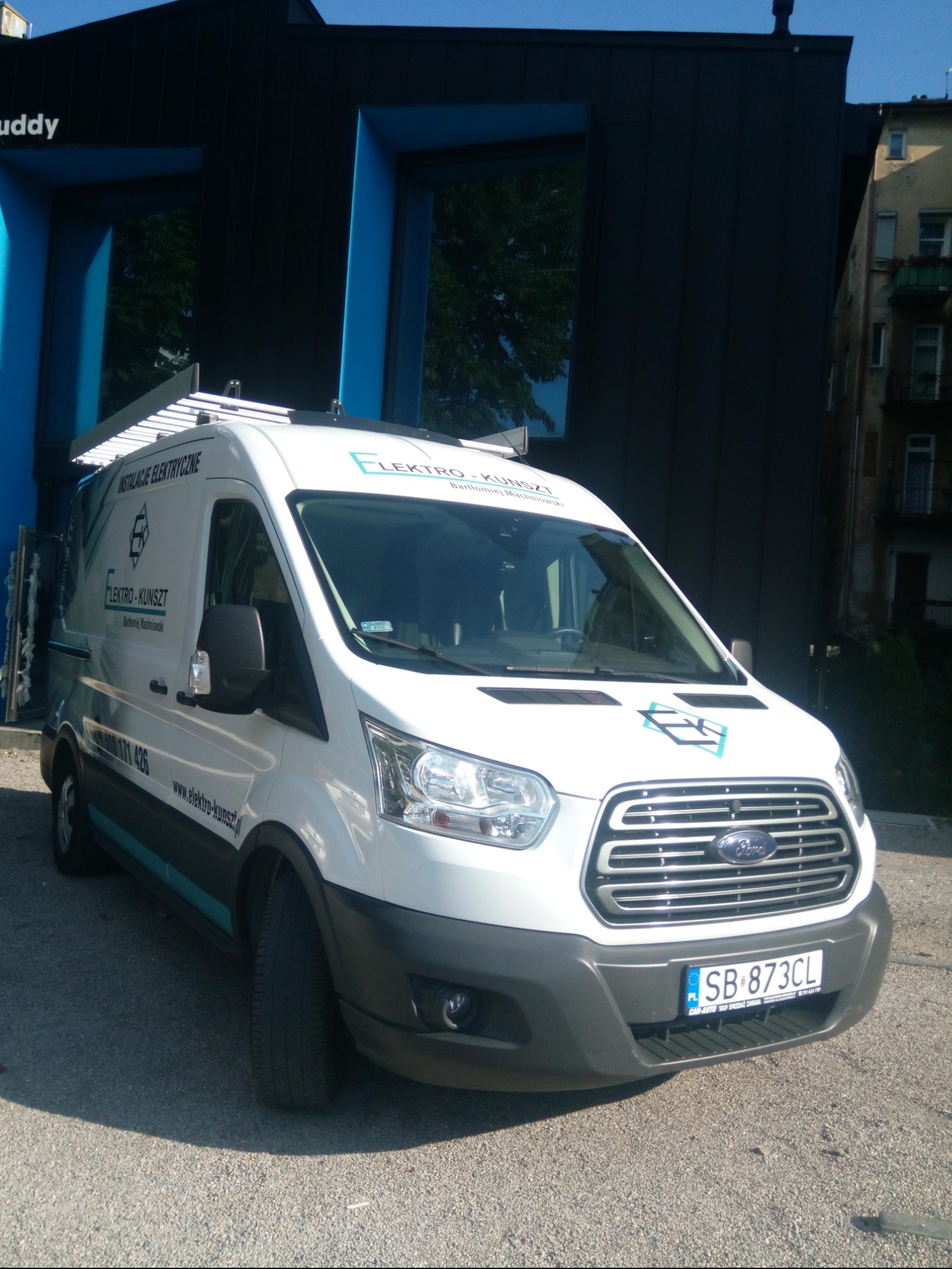 Biały samochód dostawczy firmy Elektro-Kunszt z logo i numerem telefonu na tle nowoczesnego budynku z niebieskimi akcentami.