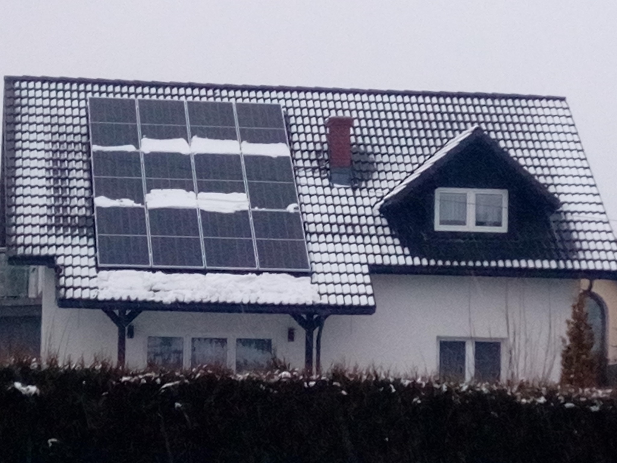 Dom jednorodzinny z panelami słonecznymi częściowo pokrytymi śniegiem na dachu w zimowy dzień.