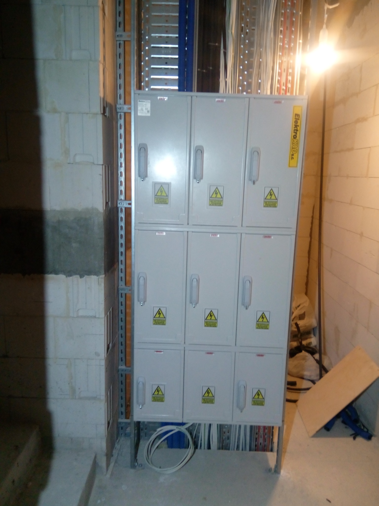 Szara, metalowa rozdzielnica elektryczna z dwunastoma drzwiczkami, każdy z symbolem ostrzegawczym, zamontowana na metalowej konstrukcji w surowym wnętrzu, widoczne przewody i elementy instalacji.