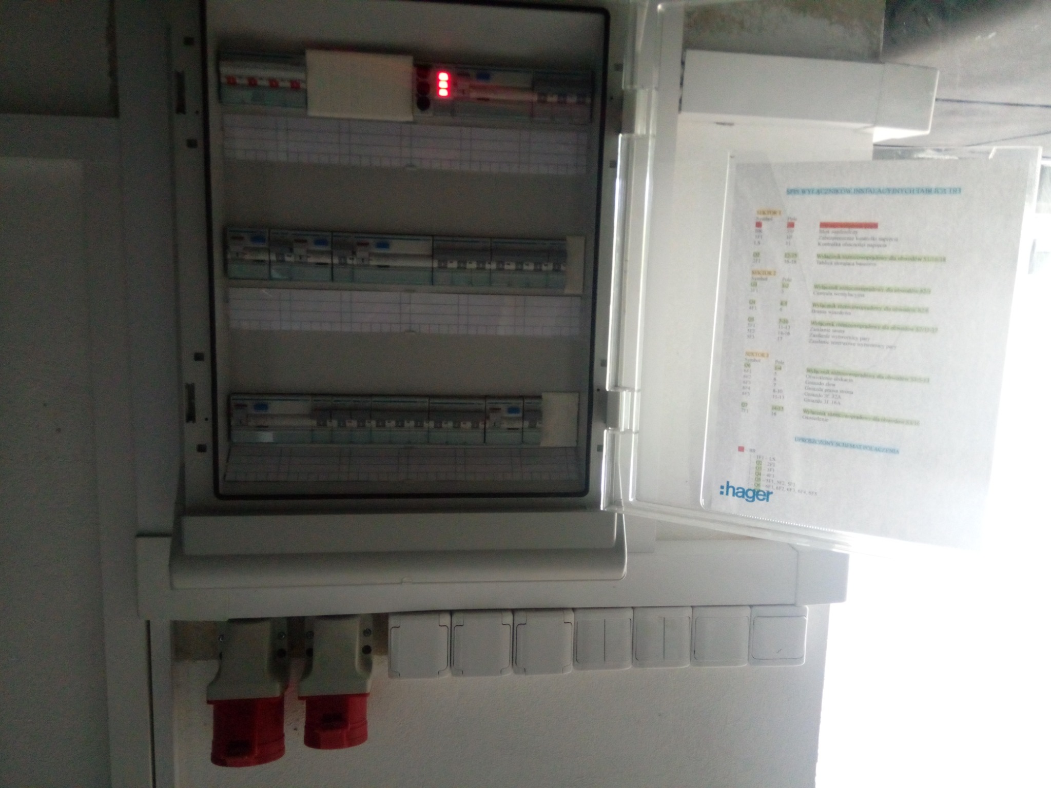 Otwarta skrzynka elektryczna z widocznymi bezpiecznikami i schematem połączeń, dwa gniazda siłowe na dole, widoczna marka Hager.