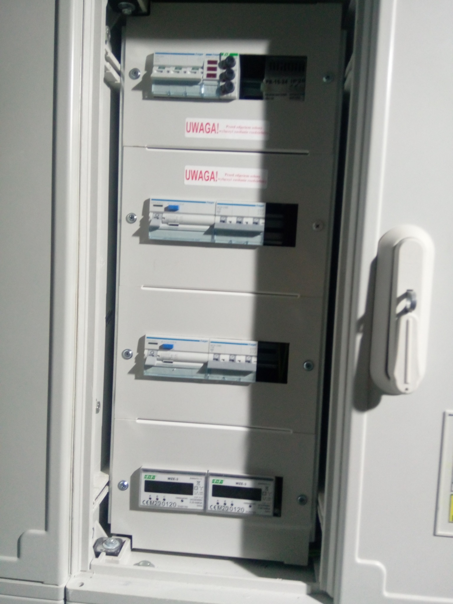 Otwarta skrzynka rozdzielcza z wyłącznikami nadprądowymi i licznikami energii elektrycznej, widoczne naklejki ostrzegawcze 'Uwaga!', całość w kolorze białym.