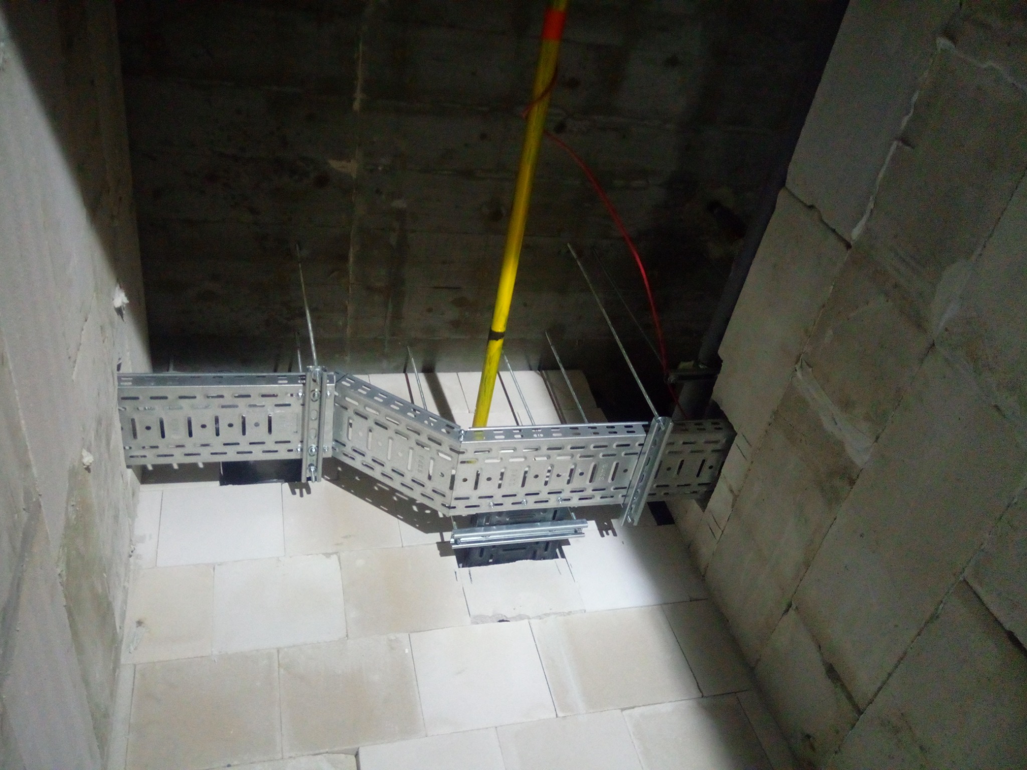 Metalowa korytka kablowa zamontowana pod betonowym sufitem, widoczne przewody w żółtej rurce i czerwony kabel, ściany z białego bloczka.