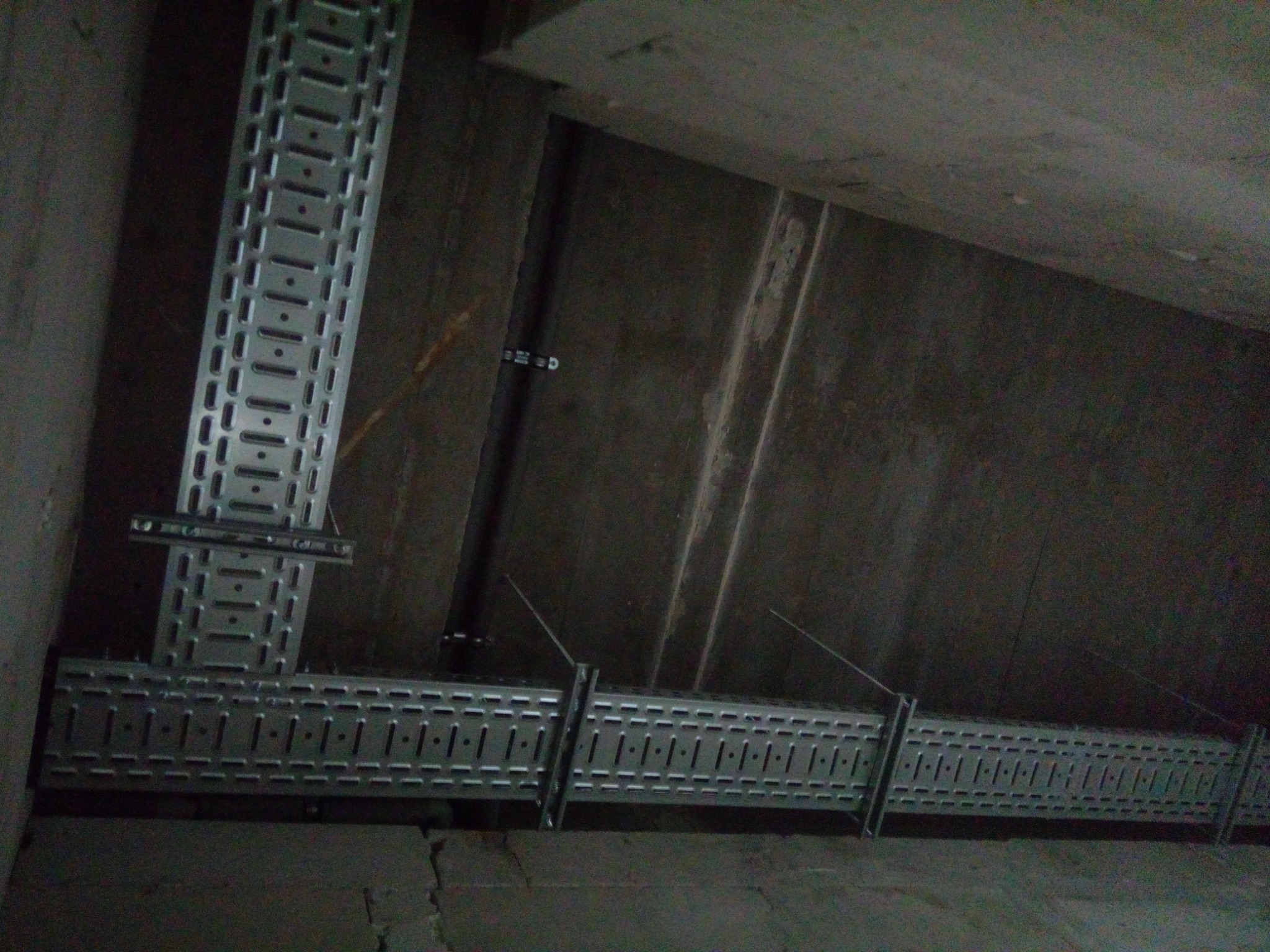 Metalowe korytka kablowe zamontowane pod sufitem w surowym pomieszczeniu, widoczne uchwyty i przewody zamocowane do ściany.