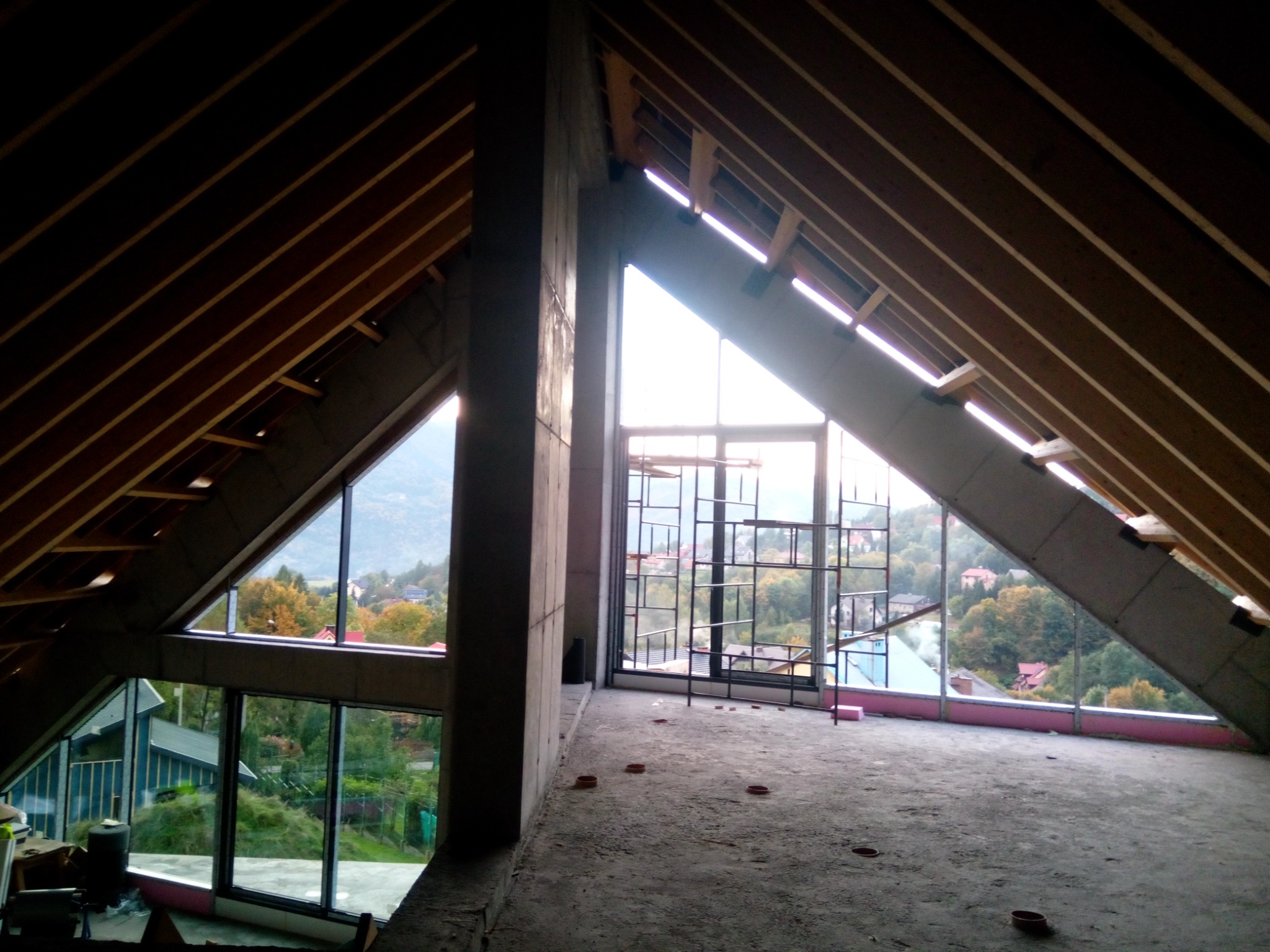 Surowy stan poddasza z drewnianą konstrukcją dachu, betonowymi filarami i dużymi oknami z widokiem na okolicę w trakcie budowy.