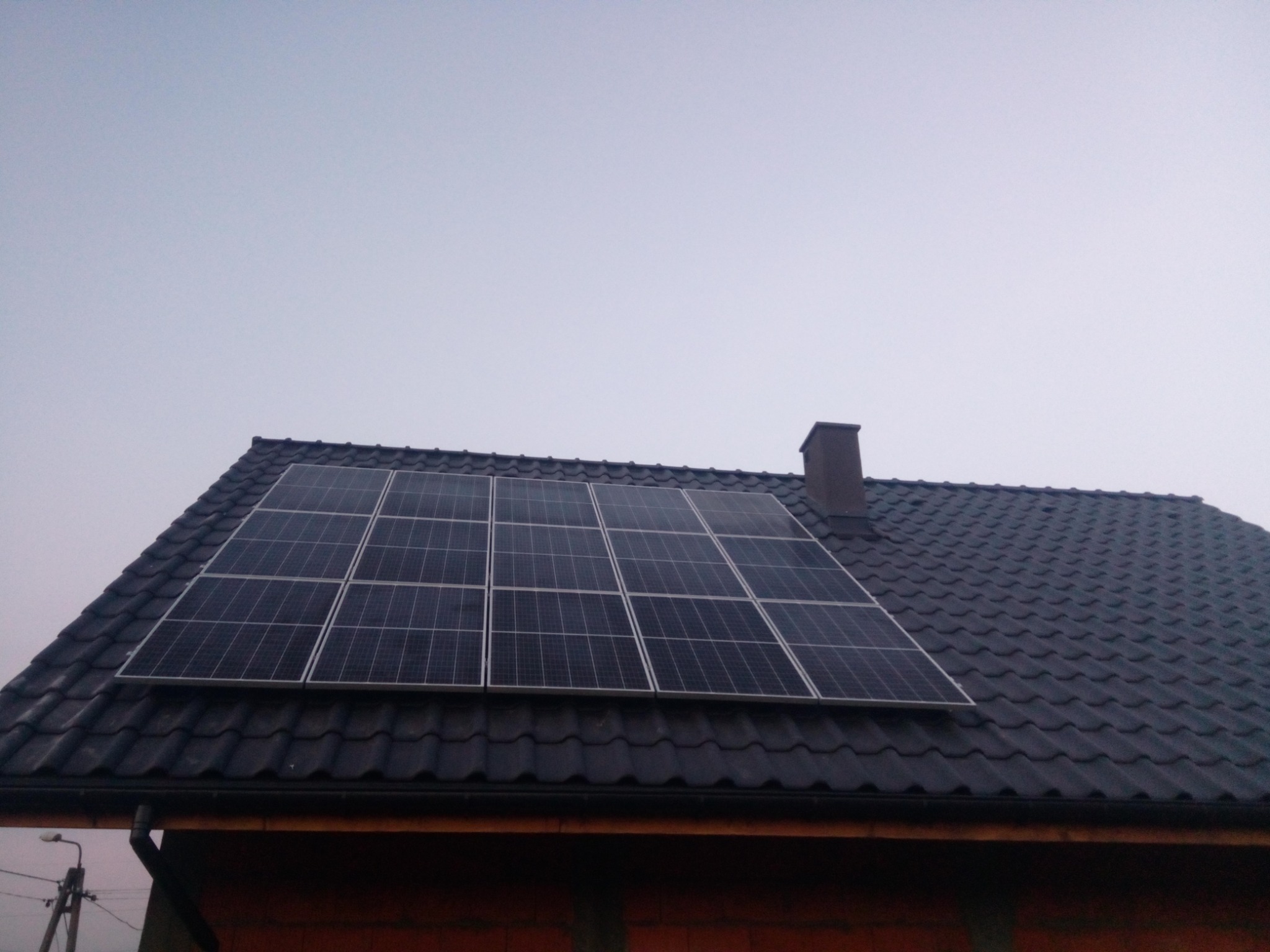 Panel fotowoltaiczny zamontowany na dachu pokrytym ciemną dachówką karpiówką, widoczny komin i fragment ściany budynku w stanie surowym.