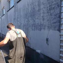 Eco Wash Service - Pracownik w czarnych wodoodpornych spodniobutach czyści elewację budynku za pomocą myjki ciśnieniowej, widoczne wyraźne różnice w kolorze elewacji przed i po czyszczeniu.