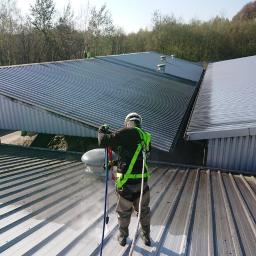Eco Wash Service - Pracownik w uprzęży, z tyłu, czyści metalowy dach przemysłowy myjką ciśnieniową, widoczne różnice w czystości dachu.