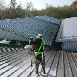 Pracownik w uprzęży, z tyłu, czyści metalowy dach przemysłowy myjką ciśnieniową, widoczne różnice w czystości dachu.