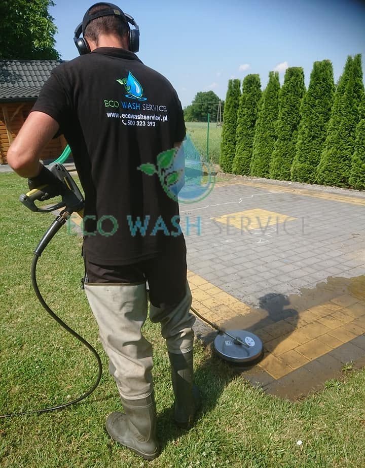 Pracownik w słuchawkach i wodoszczelnych spodniobutach czyści kostkę brukową myjką ciśnieniową rotacyjną na trawniku przed tujami.