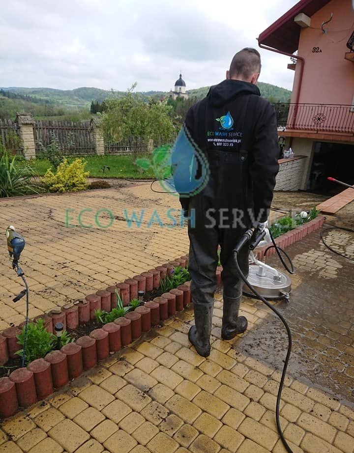 Pracownik w kombinezonie i gumowych butach czyści kostkę brukową za pomocą urządzenia wysokociśnieniowego z okrągłą dyszą, widoczny budynek z numerem 32a i kościół w tle.