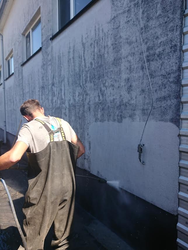 Pracownik w czarnych wodoodpornych spodniobutach czyści elewację budynku za pomocą myjki ciśnieniowej, widoczne wyraźne różnice w kolorze elewacji przed i po czyszczeniu.
