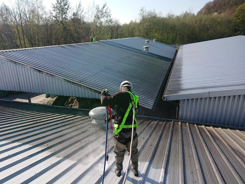 Pracownik w uprzęży, z tyłu, czyści metalowy dach przemysłowy myjką ciśnieniową, widoczne różnice w czystości dachu.