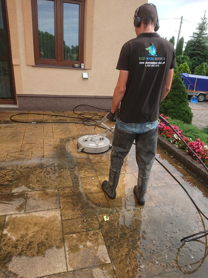 Pracownik w słuchawkach i kombinezonie czyści mokrą kostkę brukową za pomocą okrągłej myjki ciśnieniowej, widoczny budynek z oknami i kwiaty w tle, logo firmy na koszulce.