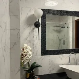 Nowoczesna łazienka z marmurowymi płytkami, umywalką w kształcie łzy, lustrem otoczonym czarną mozaiką oraz lampą ścienną z białym kloszem.