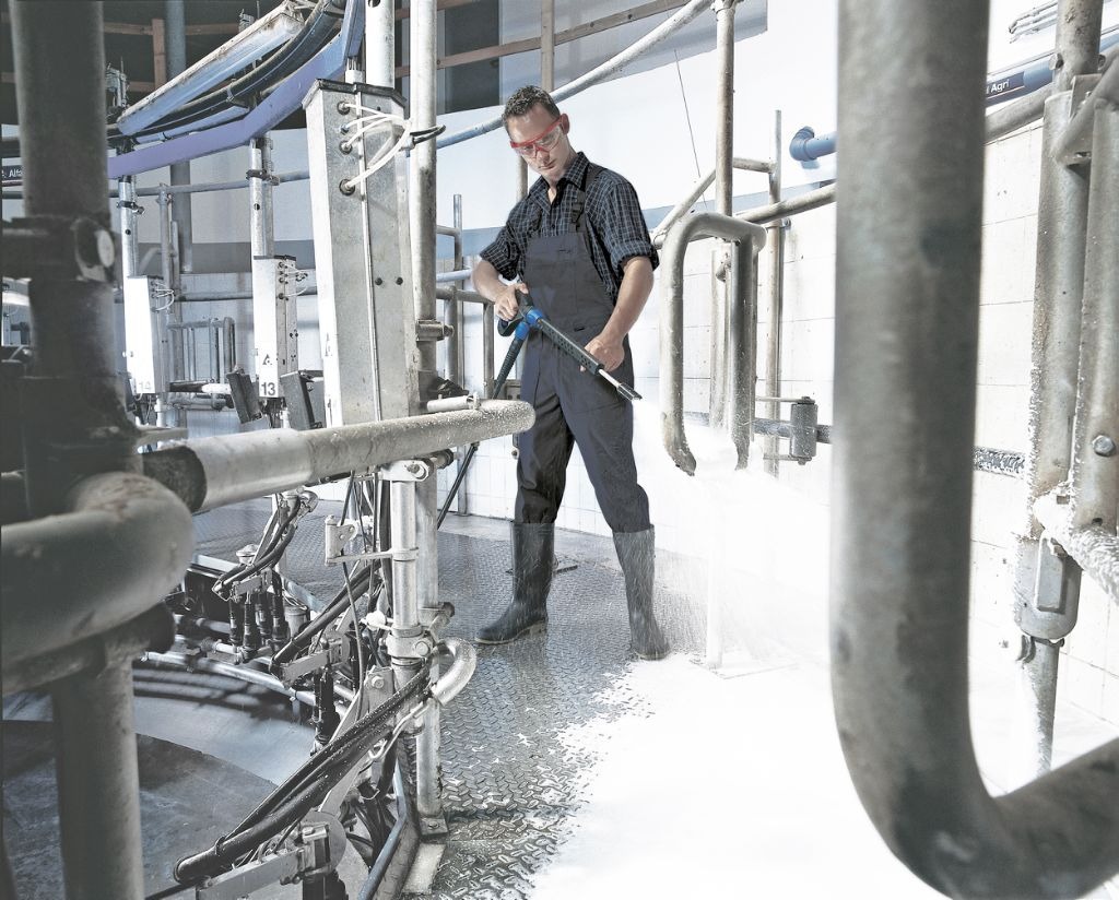 Pracownik w kombinezonie i okularach ochronnych czyści przemysłową instalację do dojenia krów za pomocą myjki ciśnieniowej, widoczne rozpryski wody i piany.