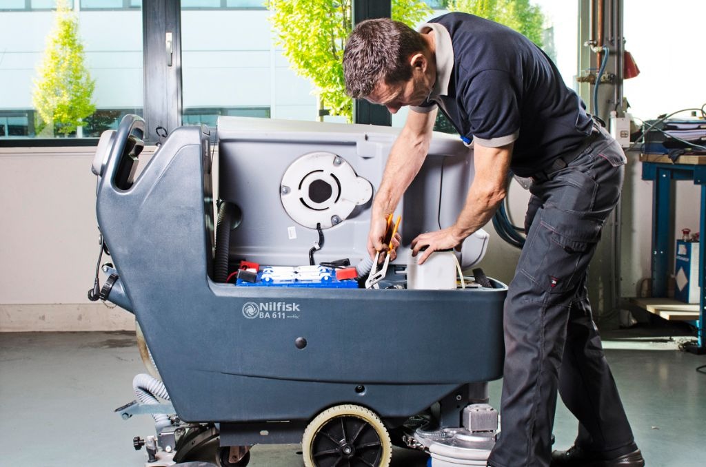 Mężczyzna w szarych spodniach roboczych naprawia lub konserwuje szorowarkę przemysłową Nilfisk BA 611, widoczne akumulatory, narzędzia i części wewnętrzne maszyny.