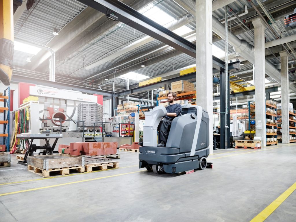 Operator na samojezdnej maszynie czyszczącej Nilfisk, przemysłowa hala z regałami i maszynami w tle, żółte linie wyznaczające trasy.