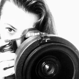 Czarno-białe zbliżenie kobiety z aparatem Nikon, widoczne oko i fragment włosów, obiektyw aparatu na pierwszym planie.