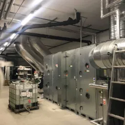 Instalacja wentylacji przemysłowej z centralami wentylacyjnymi, rurami wentylacyjnymi pod sufitem i zbiornikiem na płyn na palecie, drabina przystawna z boku.