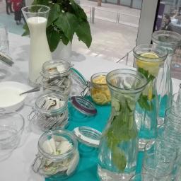 Mm Catering Marcin Burzak - Stół z przygotowanym bufetem napojów: karafki z wodą z cytryną i miętą, dzbanek z mlekiem, słoiki z herbatami i dodatkami, ułożone szklanki i łyżeczki.