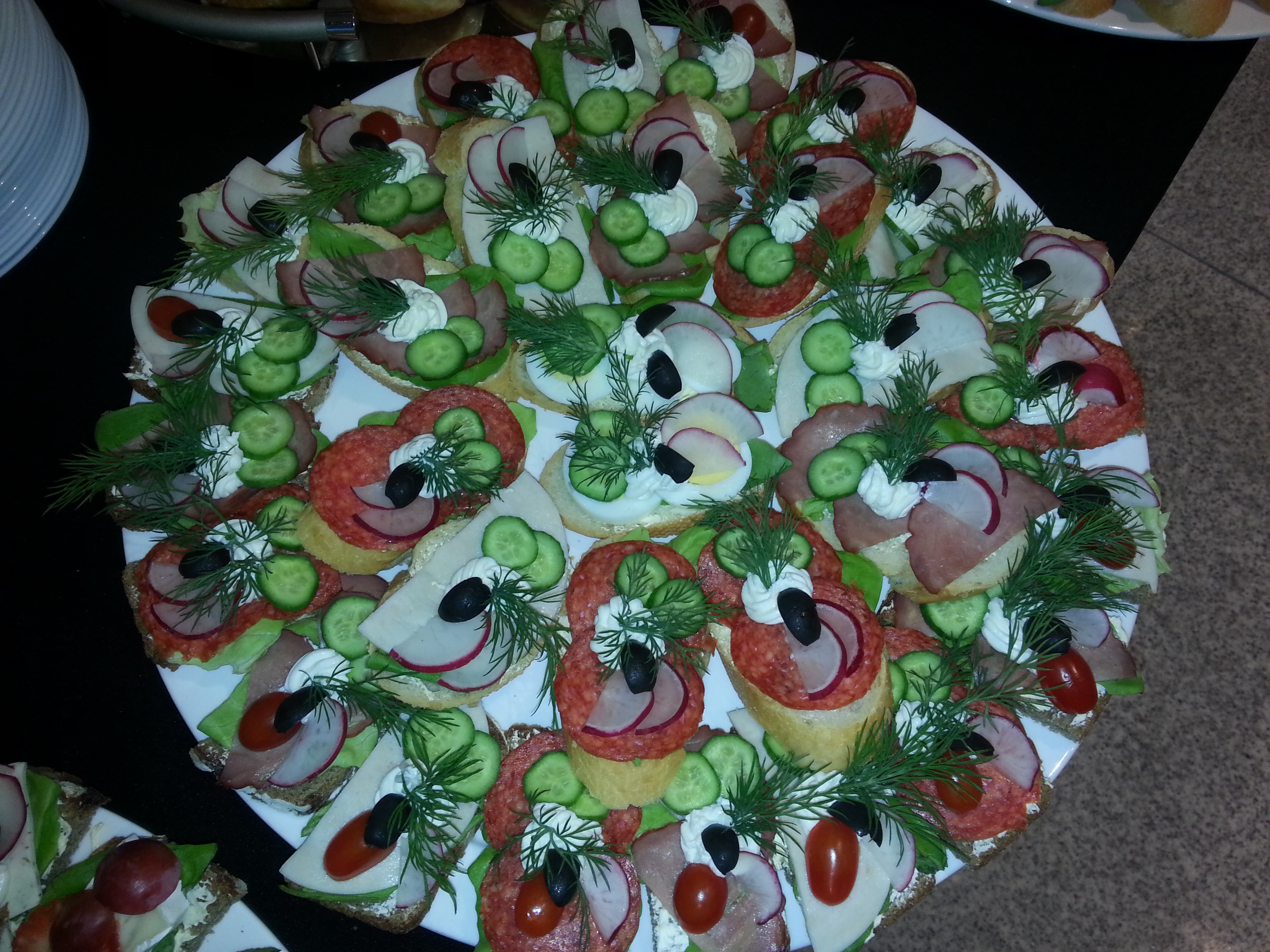 Duży biały talerz z różnorodnymi, bogato zdobionymi kanapkami bankietowymi, z salami, szynką, rzodkiewką, ogórkiem, oliwkami i koperkiem, ułożonymi na jasnym pieczywie.