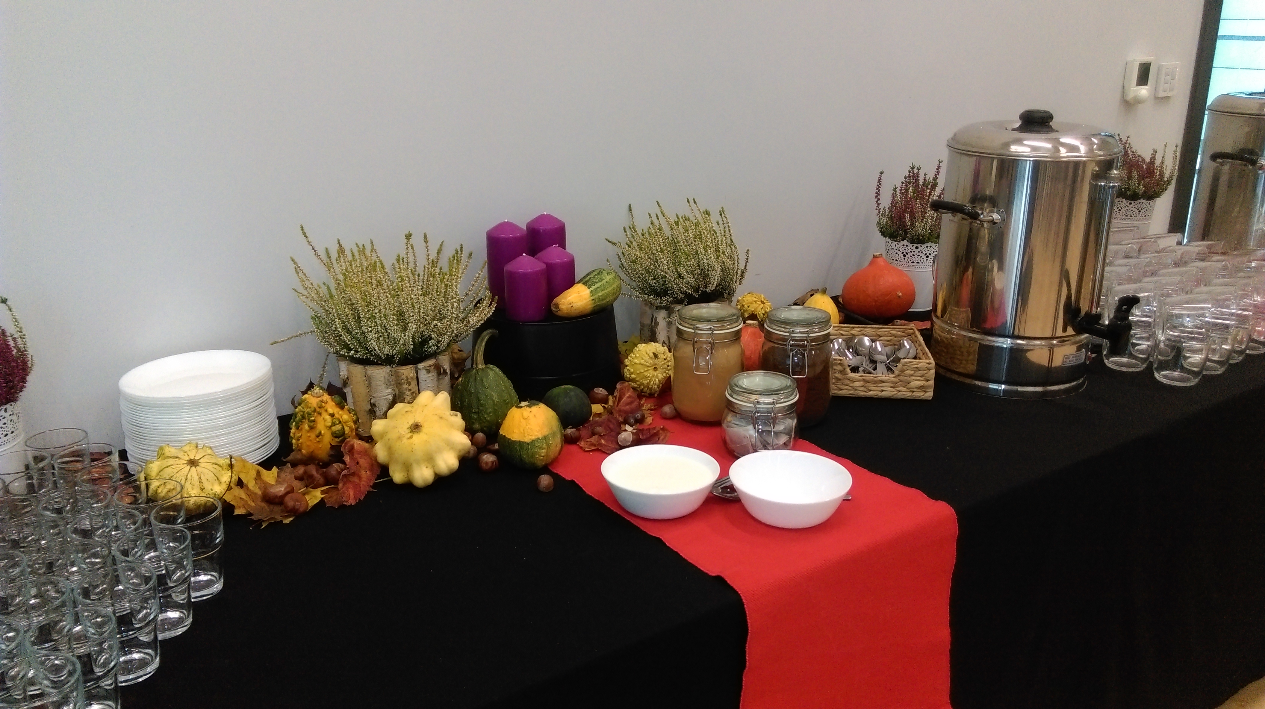 Jesienna dekoracja stołu cateringowego z dyniami ozdobnymi, wrzosami, świecami i naczyniami z zupą krem, obok zapas czystych szklanek i termos z wrzątkiem.