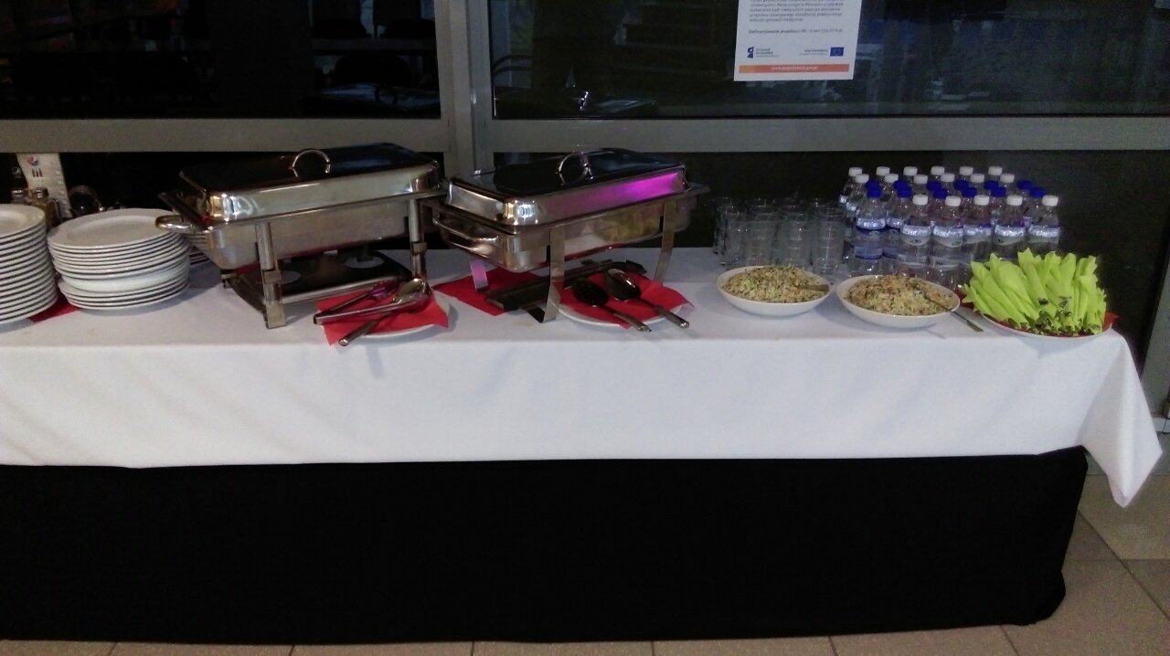 Bufet z dwoma podgrzewaczami, stosem talerzy, miskami sałatek, butelkami wody i serwetkami ułożonymi na białym obrusie z czarnym wykończeniem.