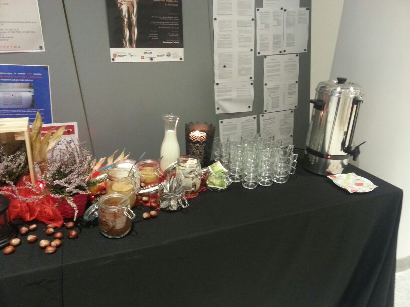 Stół cateringowy z dekoracjami: słoiczki z zawartością, dzbanek z mlekiem, orzechy laskowe, szklanki ułożone w stosy, termos z napojem, dekoracje kwiatowe z czerwonymi akcentami.