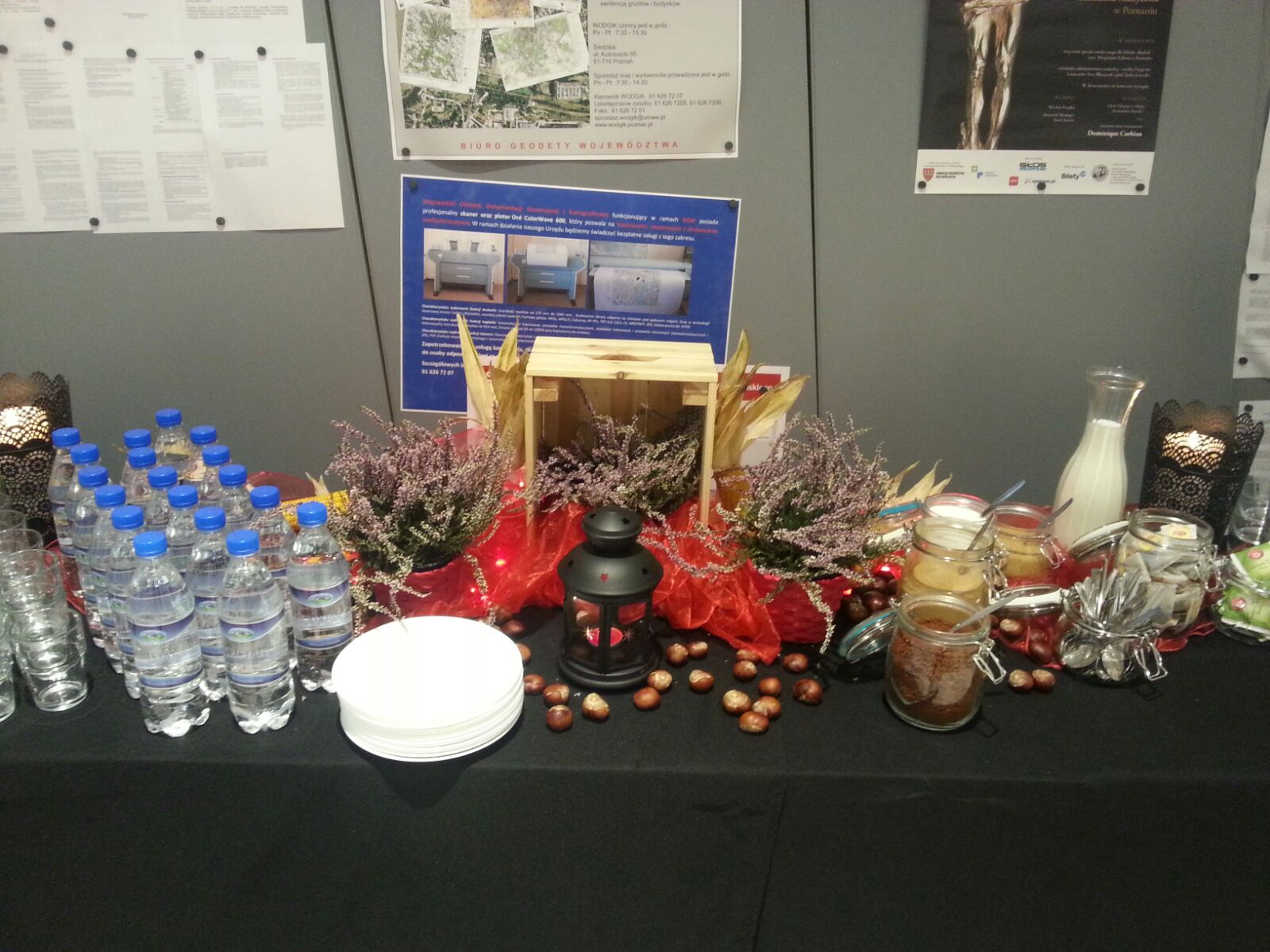 Stół z dekoracjami na firmowej imprezie: woda butelkowana, stos talerzyków, słoiki z zawartością i wrzosowe dekoracje w drewnianej skrzynce, oświetlone lampionem.