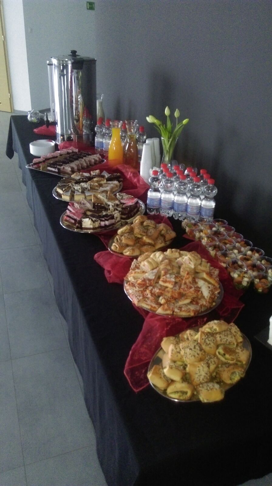 Stół cateringowy z różnorodnymi przekąskami: ciasta, mini pizze, roladki z farszem, sałatki w kubeczkach, woda butelkowana, sok pomarańczowy, termos ze stali nierdzewnej i dekoracyjne tulipany.