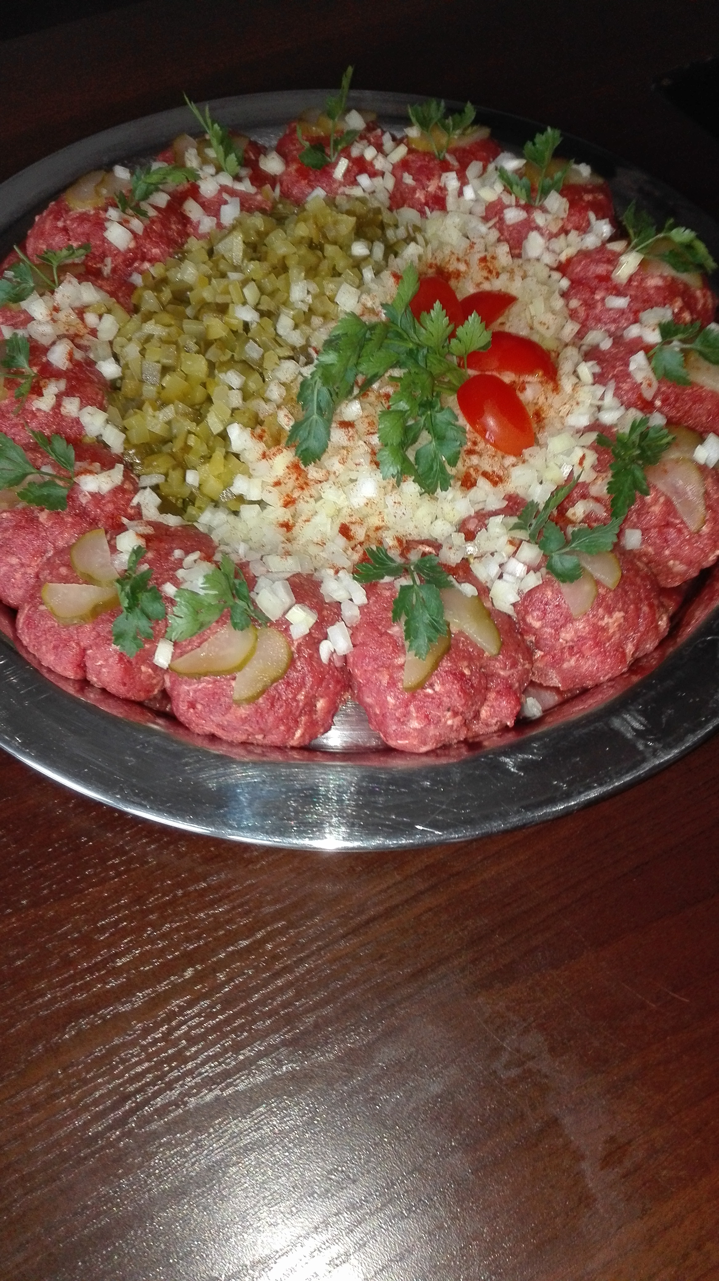 Tatar wołowy na srebrnej tacy, udekorowany posiekaną cebulą, ogórkiem kiszonym, natką pietruszki i papryką w proszku, z pomidorami na środku.