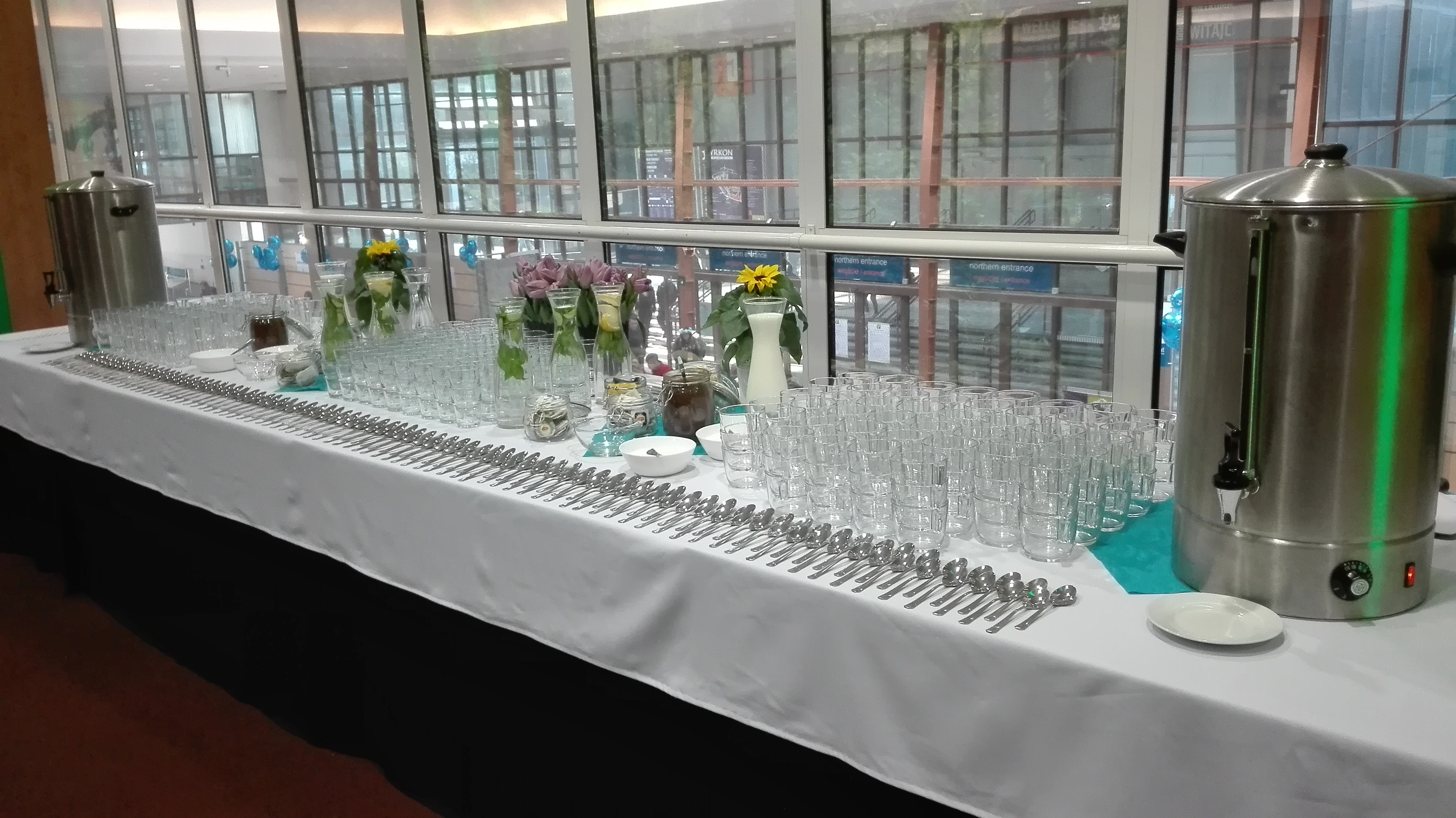 Stół z białym obrusem zastawiony szklankami, łyżeczkami, dzbankami z wodą i kwiatami, oraz dwoma metalowymi podgrzewaczami do wody na tle przeszklonej ściany.