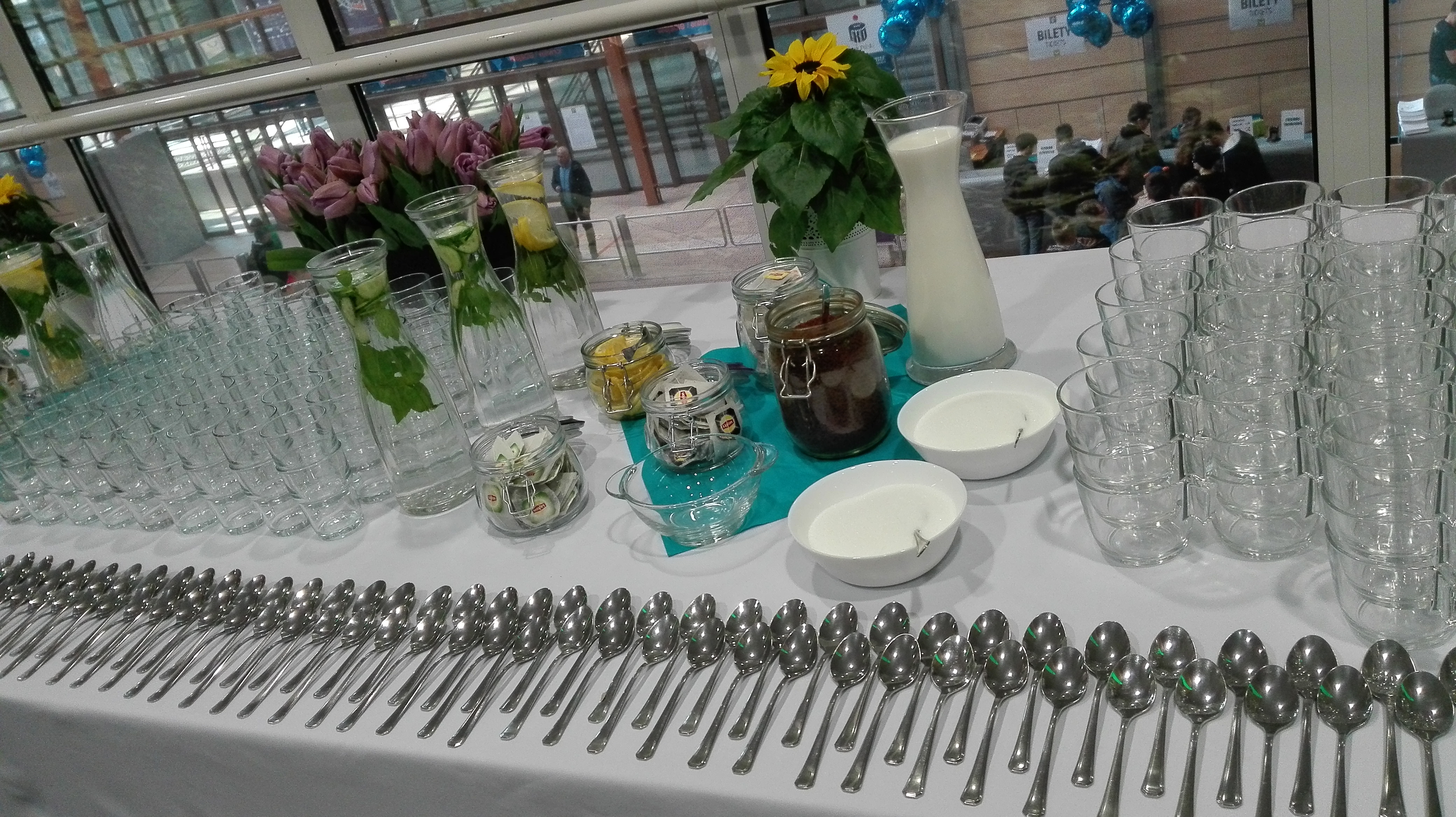 Stół z przygotowanym bufetem: dzbanki z wodą smakową, słoiki z herbatą i dżemem, miseczki z cukrem, stosy szklanek i rzędy srebrnych łyżeczek.