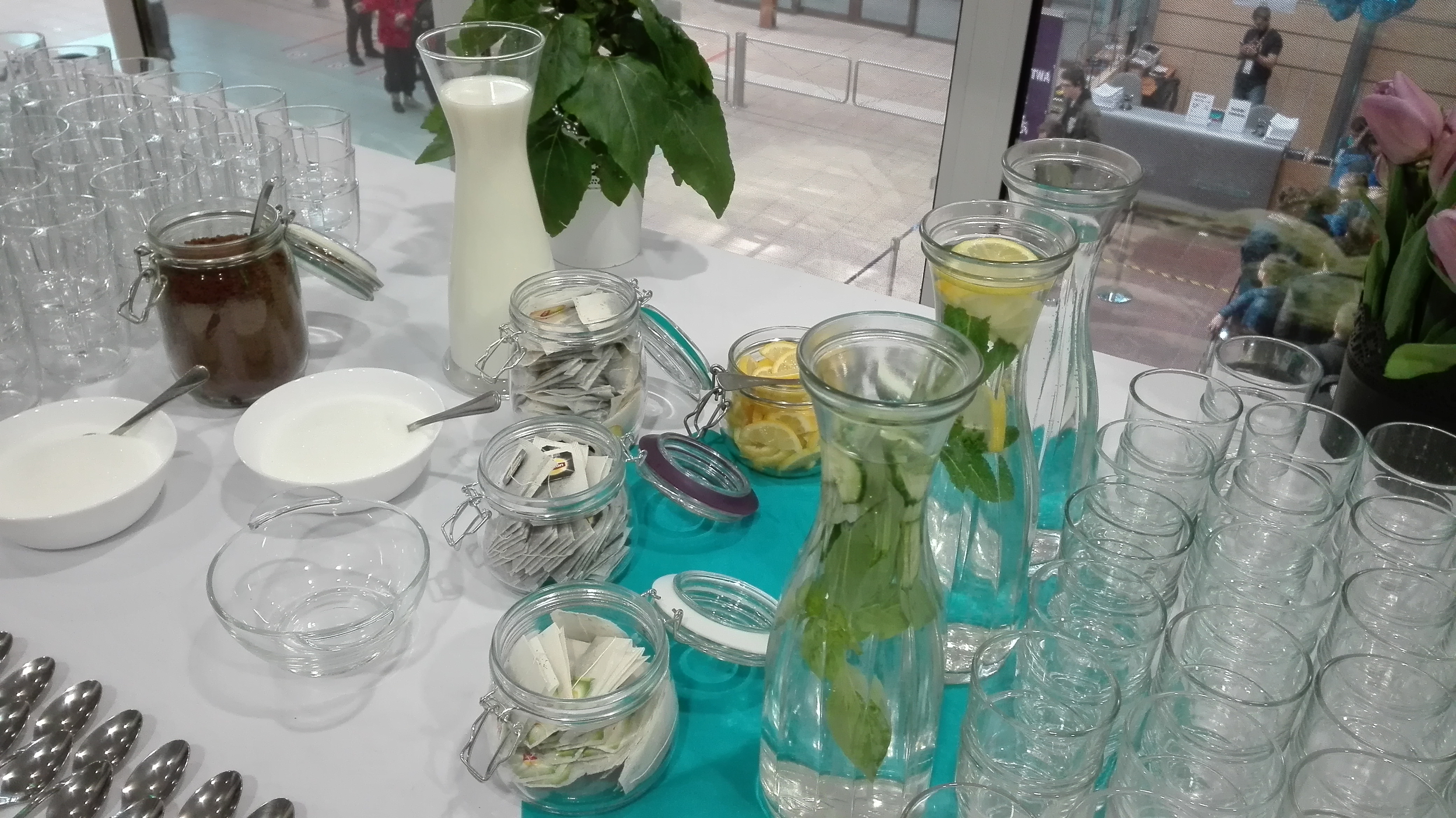 Stół z przygotowanym bufetem napojów: karafki z wodą z cytryną i miętą, dzbanek z mlekiem, słoiki z herbatami i dodatkami, ułożone szklanki i łyżeczki.