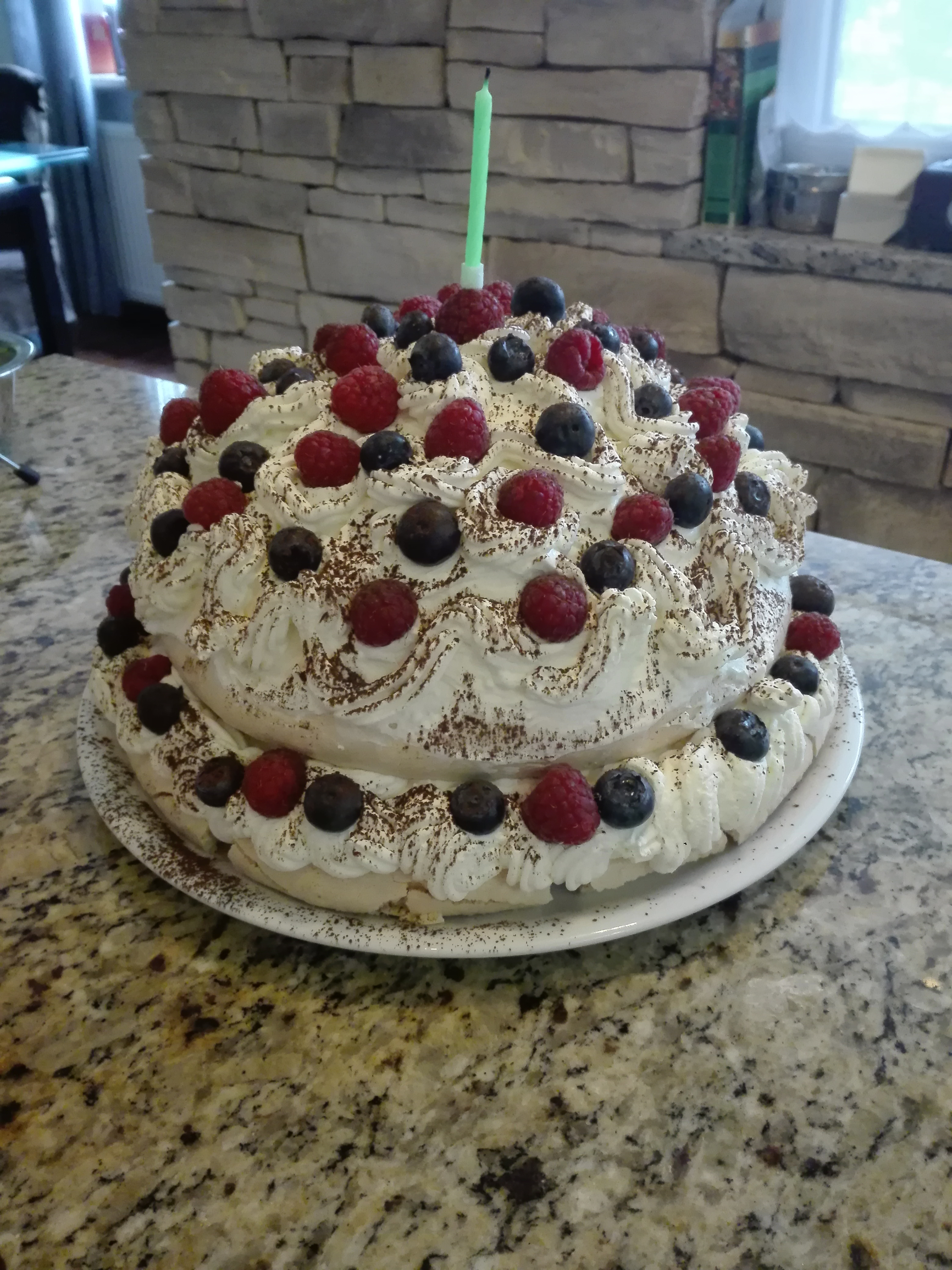 Dwupiętrowy tort Pavlova z bitą śmietaną, malinami, borówkami i posypką kakaową, zwieńczony zieloną świeczką, na białym talerzu i granitowym blacie.