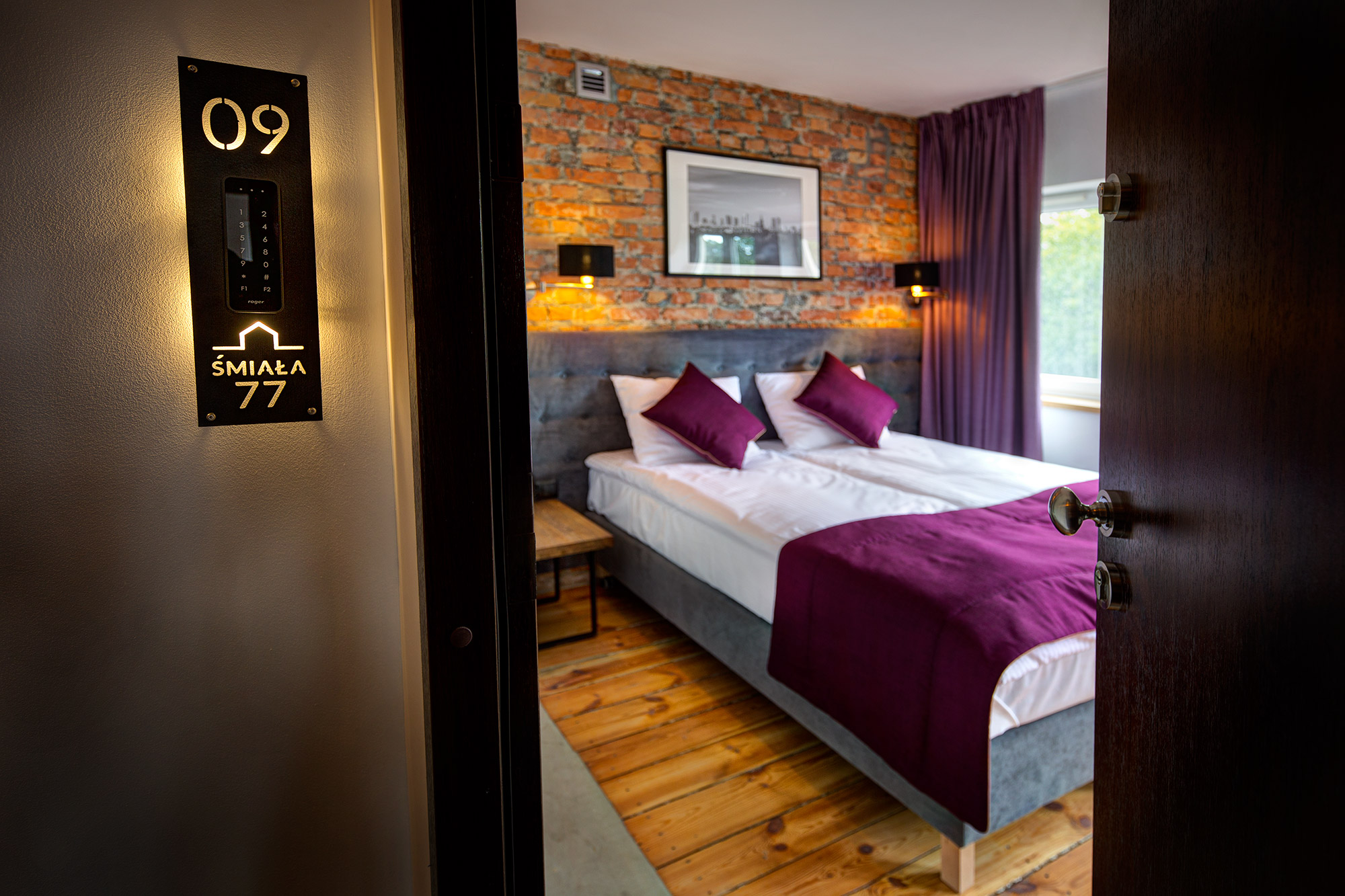 Wnętrze pokoju hotelowego widziane przez uchylone drzwi, z widocznym panelem z numerem pokoju '09 Śmiała 77' z podświetleniem LED, łóżkiem z purpurowymi poduszkami i ceglaną ścianą w tle.