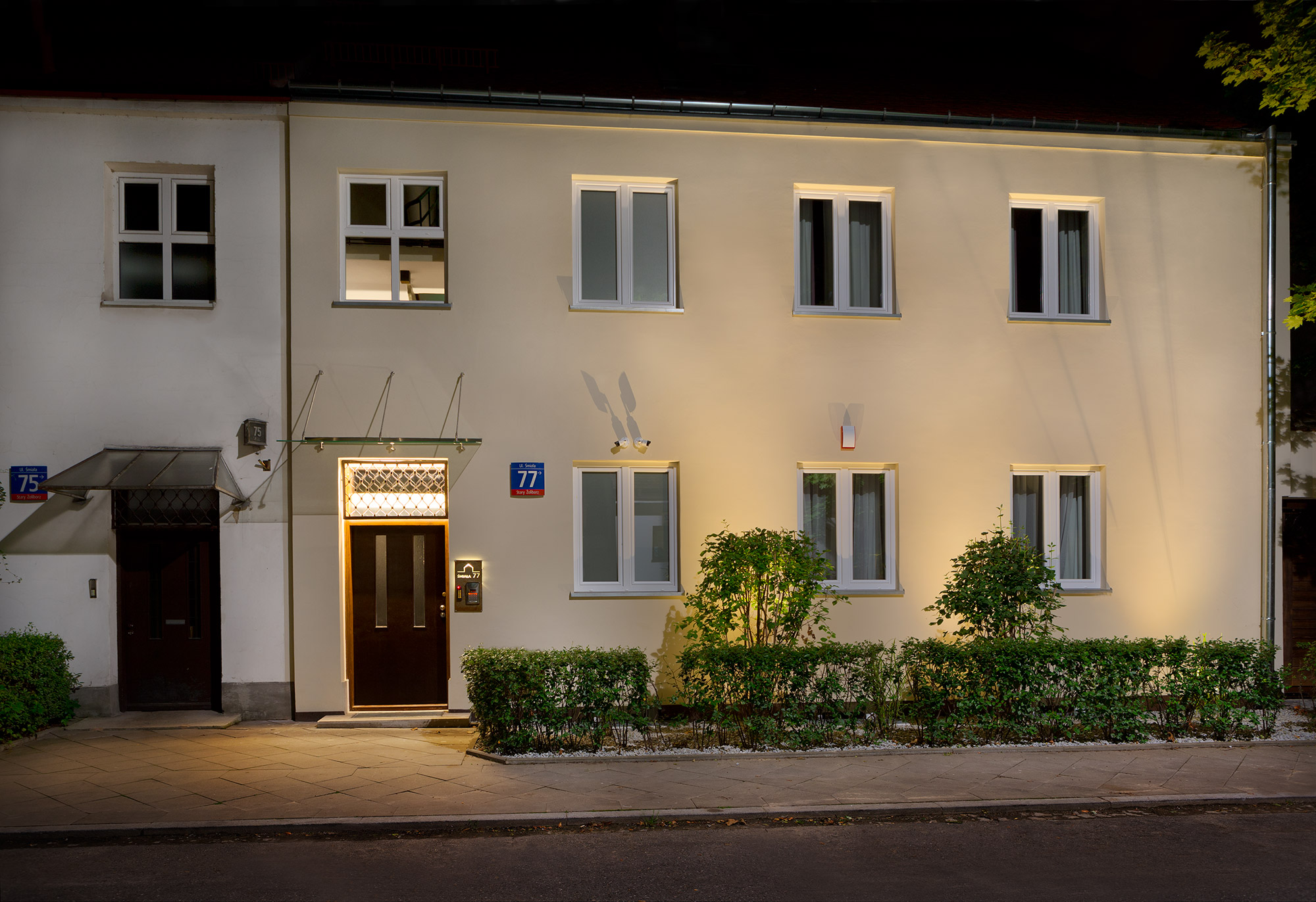 Elewacja budynku oświetlona nocą, z numerem 77 na niebieskiej tabliczce, ciemne drzwi wejściowe podświetlone ciepłym światłem, niski żywopłot przed budynkiem.