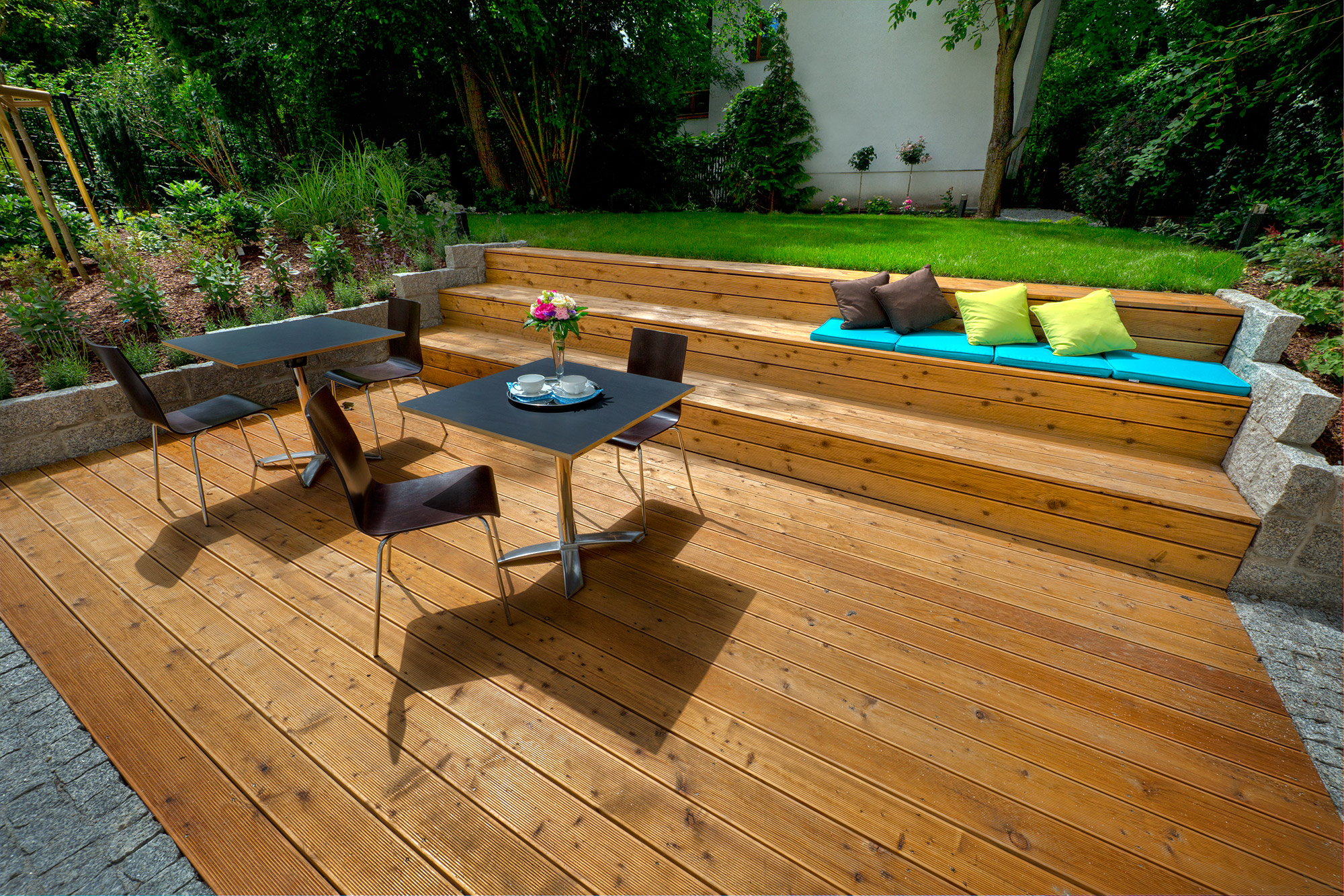 Drewniany taras z wielopoziomową konstrukcją, siedziskiem z poduszkami i stolikami kawowymi, otoczony zielenią.