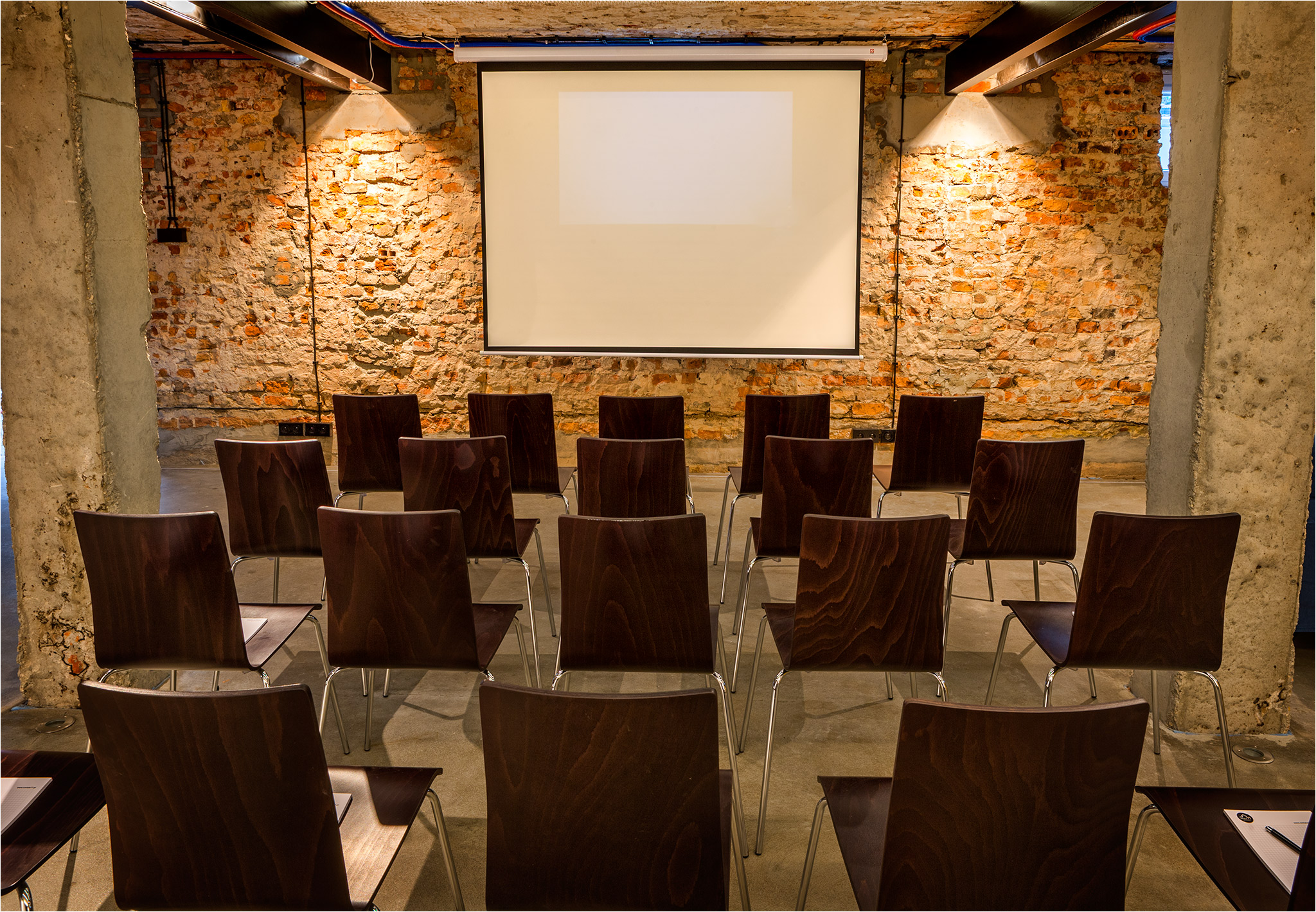 Sala konferencyjna z rzędami ciemnobrązowych krzeseł ustawionych przed ekranem projekcyjnym, surowa ceglana ściana w tle, oświetlenie punktowe.