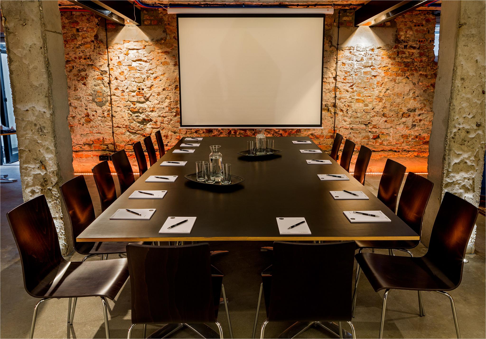 Sala konferencyjna z długim stołem z ciemnego drewna, krzesłami z giętego drewna, szklankami i karafkami na stole, notesami i długopisami dla uczestników, oraz ekranem projekcyjnym na tle ceglanej...