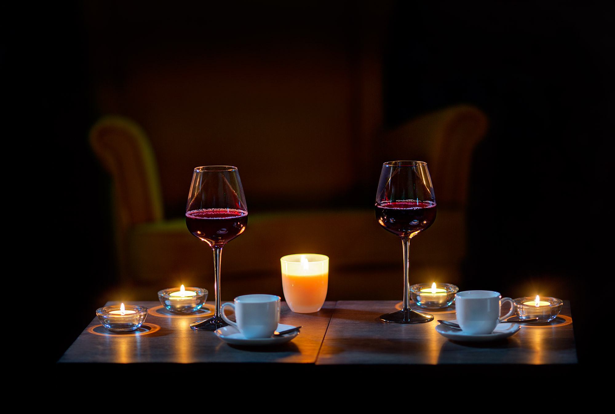Romantyczna kolacja w Warszawie: Dwa kieliszki czerwonego wina i filiżanki kawy przy świecach na drewnianym stole, w tle rozmyte żółte krzesło.