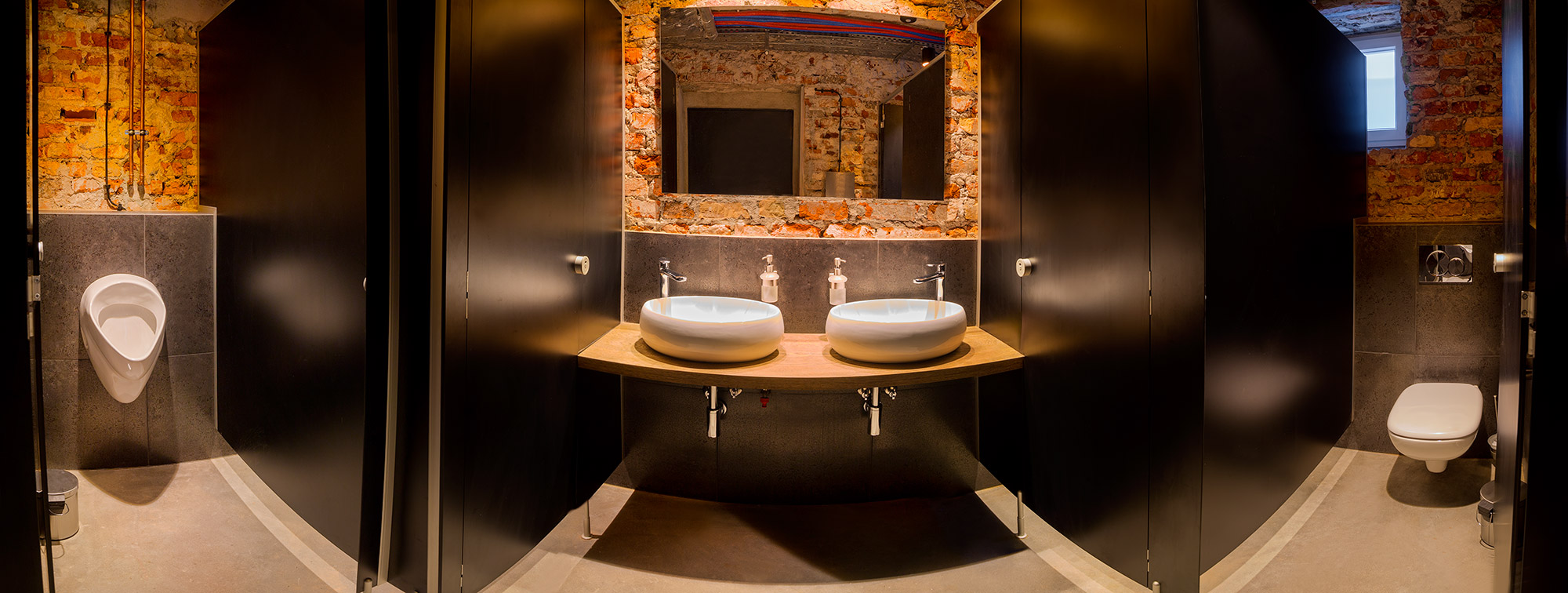 Nowoczesna toaleta publiczna z dwoma umywalkami w stylu loft, odsłonięta cegła, kabiny z czarnymi drzwiami.