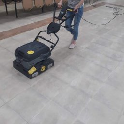 Czysty Dom - Osoba obsługuje profesjonalną maszynę do czyszczenia podłóg z szarych płytek w pomieszczeniu z krzesłami ustawionymi na stołach.