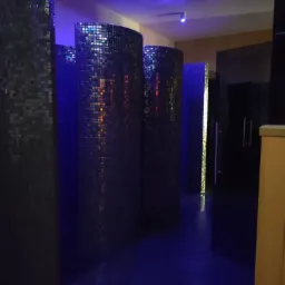 Wnętrze łazienki z cylindrycznymi ściankami prysznicowymi wyłożonymi mozaiką w odcieniach szarości i błękitu, oświetlone niebieskim światłem punktowym.