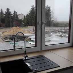 Czysty Dom - Kuchenny zlew z czarnego granitu z widokiem na plac budowy za oknem, z hałdą piasku i błotem po deszczu.
