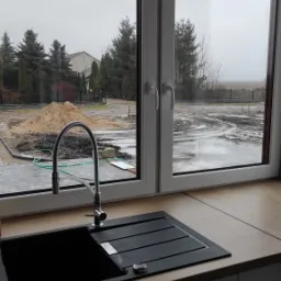 Kuchenny zlew z czarnego granitu z widokiem na plac budowy za oknem, z hałdą piasku i błotem po deszczu.