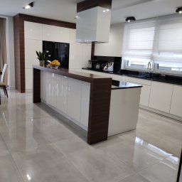 Czysty Dom - Nowoczesna, minimalistyczna kuchnia z wyspą, białymi szafkami i drewnianymi akcentami, widok na błyszczące, jasne płytki podłogowe.